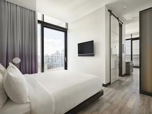 A modern hotel room with a large bed, a TV mounted on the wall, and a city view through the window.