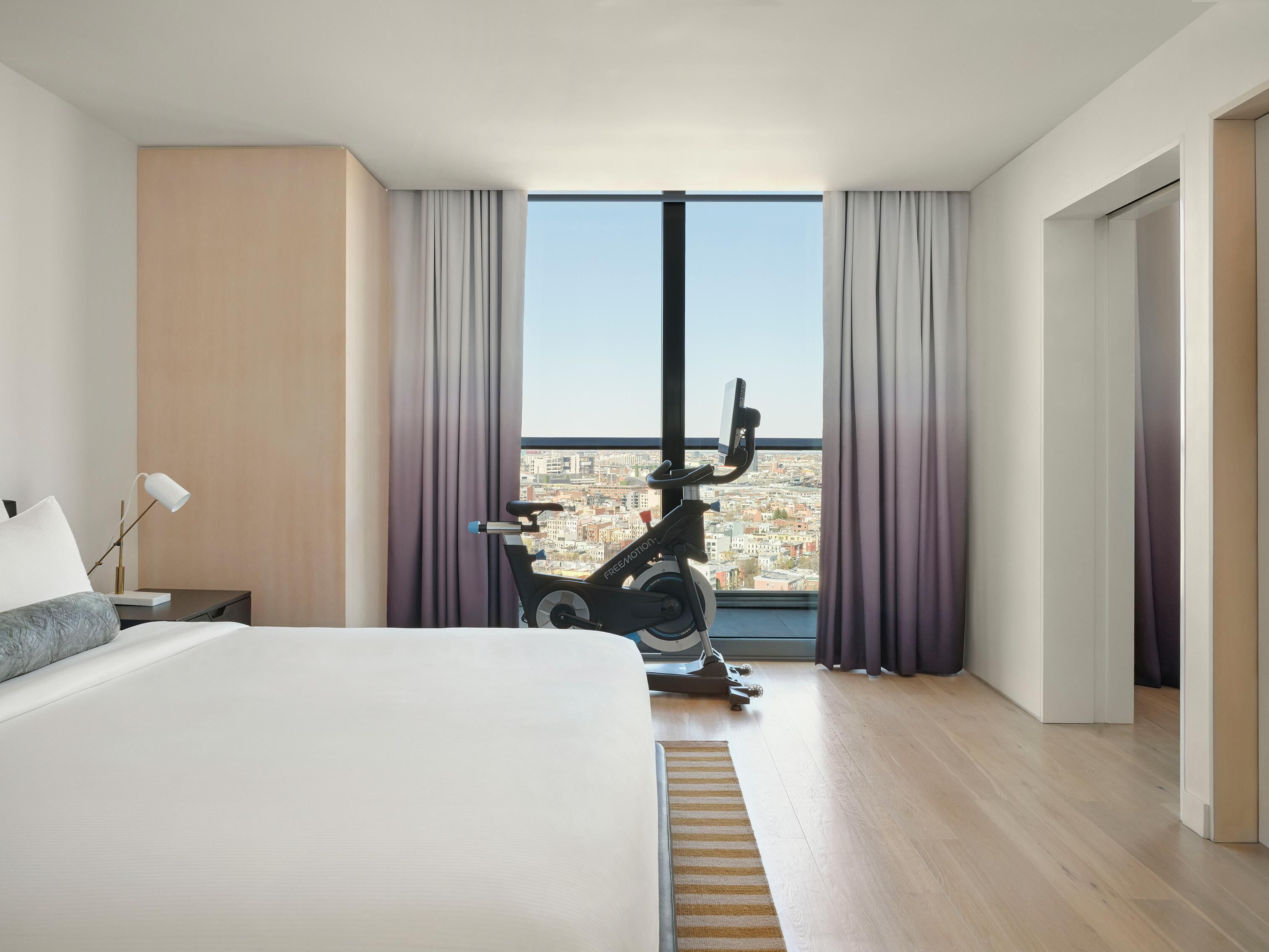 A modern hotel room with a large bed, a standing exercise bike, and a city view through large windows.