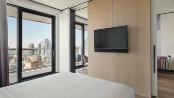 A modern hotel room with a large bed, a flat-screen TV mounted on the wall, and a city view through large windows.