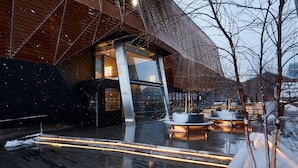 En moderne bygning med en geometrisk facade, store vinduer og udendørs siddeområder med oplyste borde.
