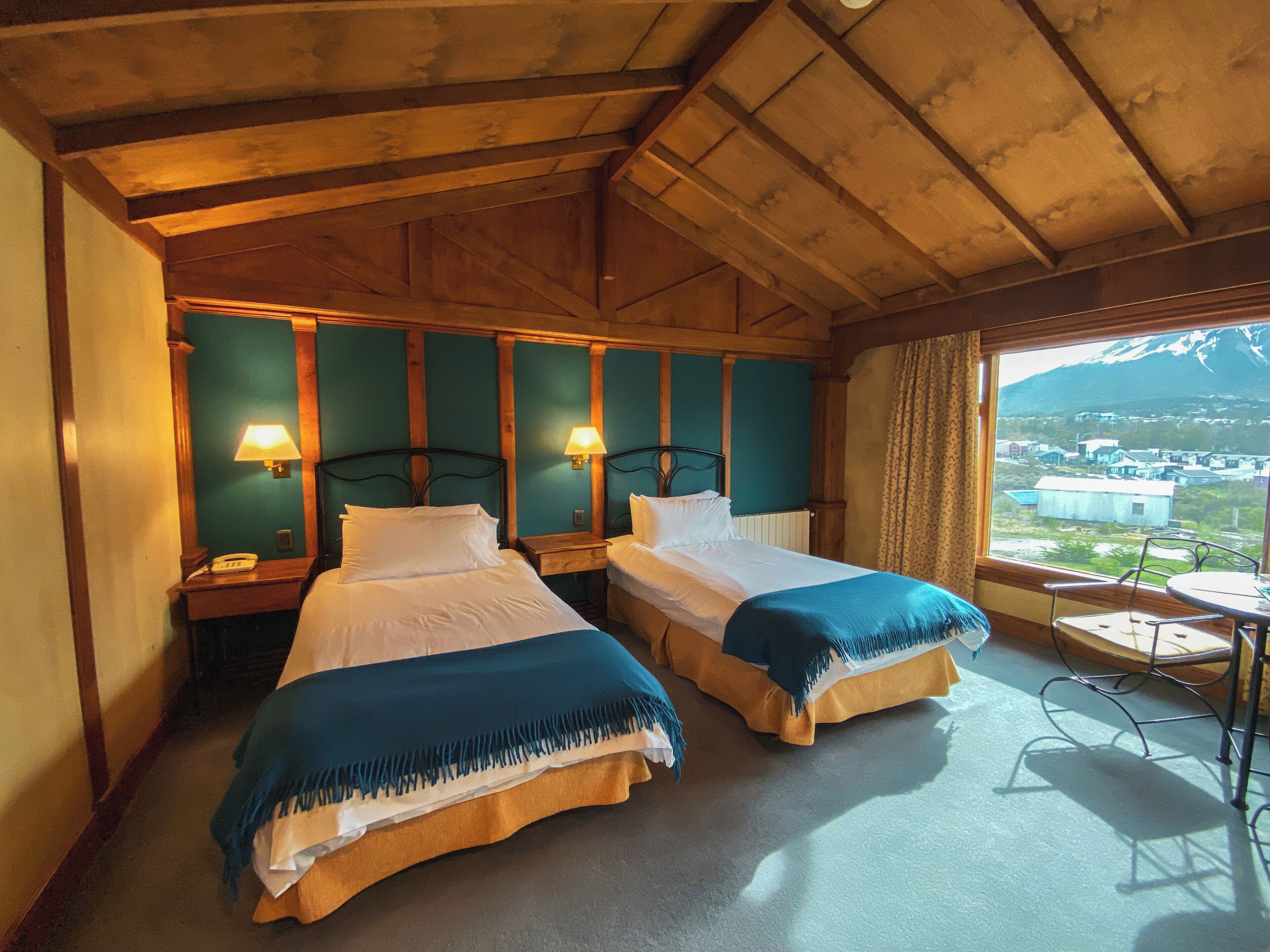 A room with two beds, wooden walls, a window with a view, and a small table.
