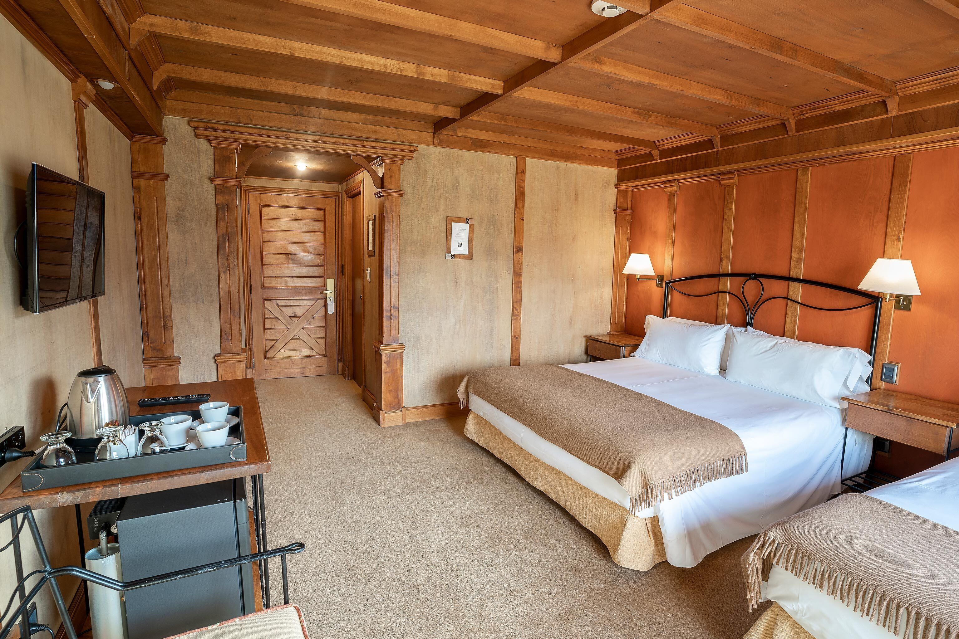 Quarto de hotel com teto de madeira, cama com roupa de cama branca, duas mesas de cabeceira com luminárias, escrivaninha com cafeteira e televisão na parede.