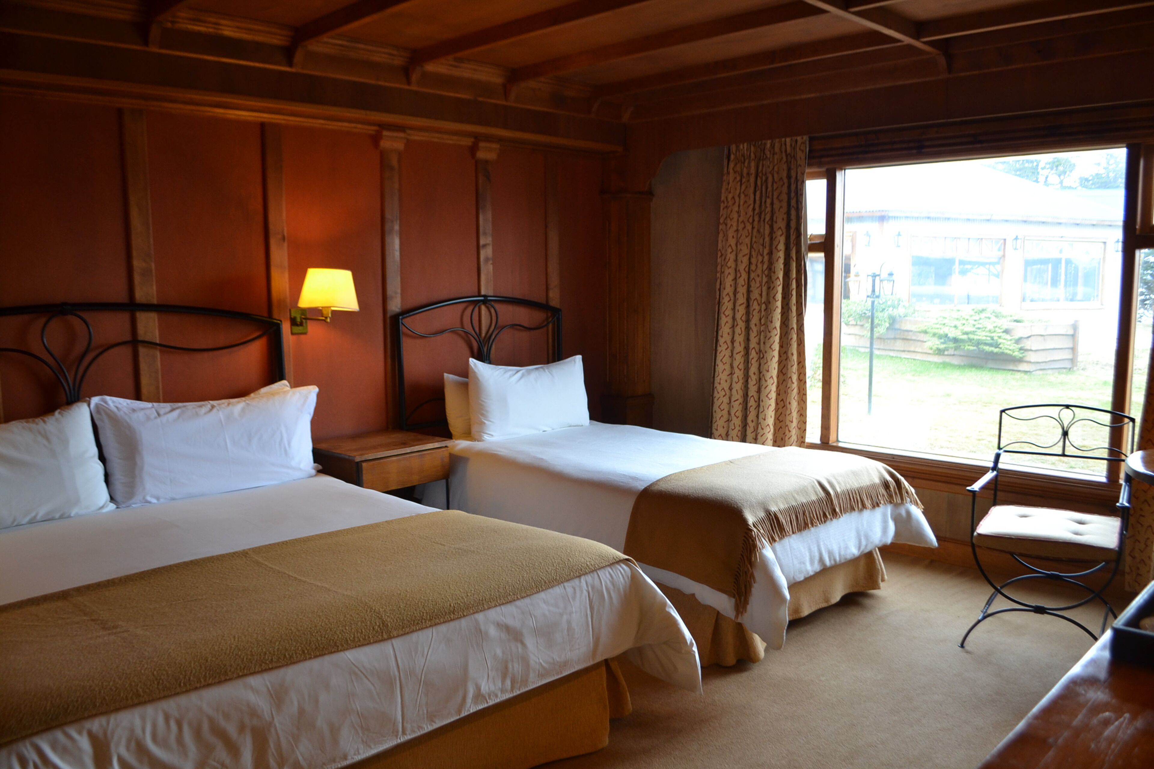 A hotel room with two beds, a nightstand, a lamp, a chair, and a window with a view of the outside.