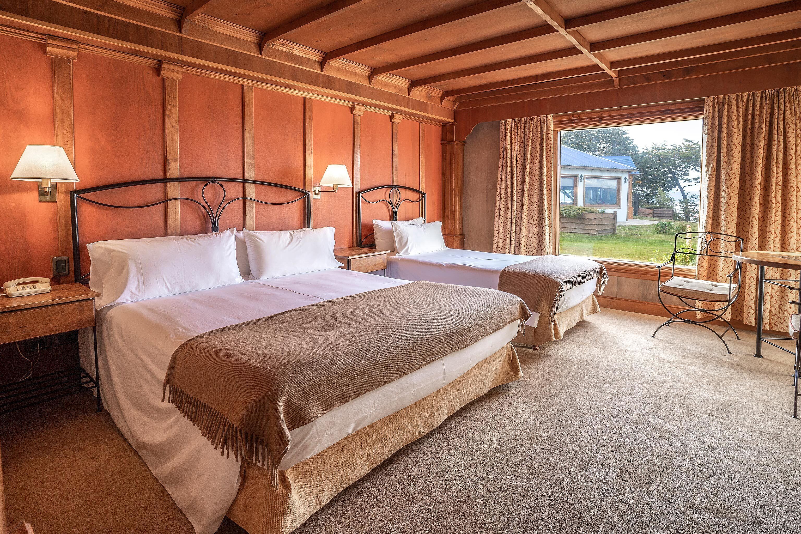 A hotel room with two beds, wooden paneling, a desk with a lamp, and a view of a house and garden.
