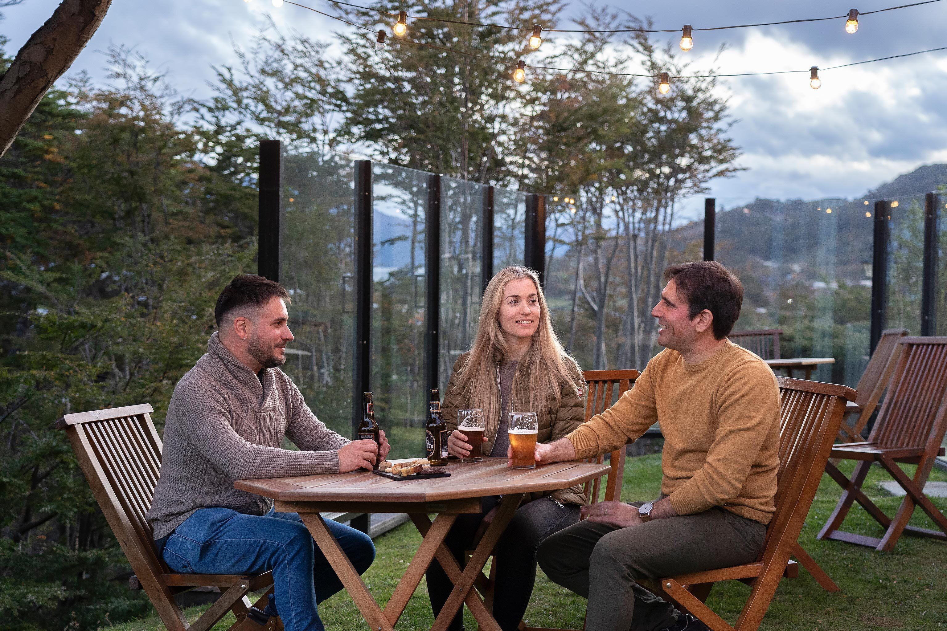 Three people seated at a wooden table outdoors with string lights above, surrounded by wooden chairs and a glass partition.