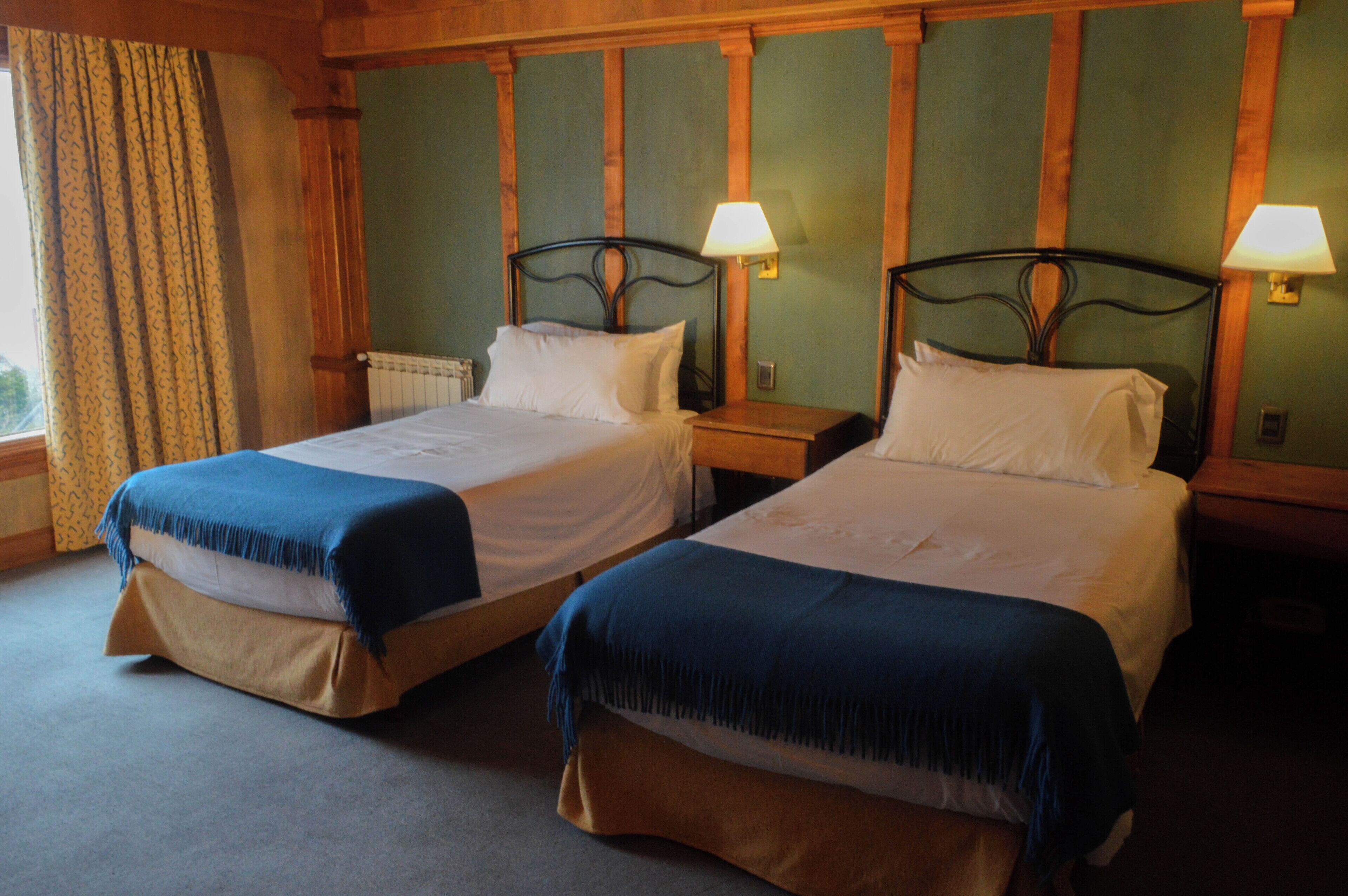 Habitación de hotel con dos camas, cada una con cubrecama azul y almohadas blancas. Hay dos mesitas de noche con lámparas. La habitación tiene paneles de madera en las paredes y una ventana con cortinas.