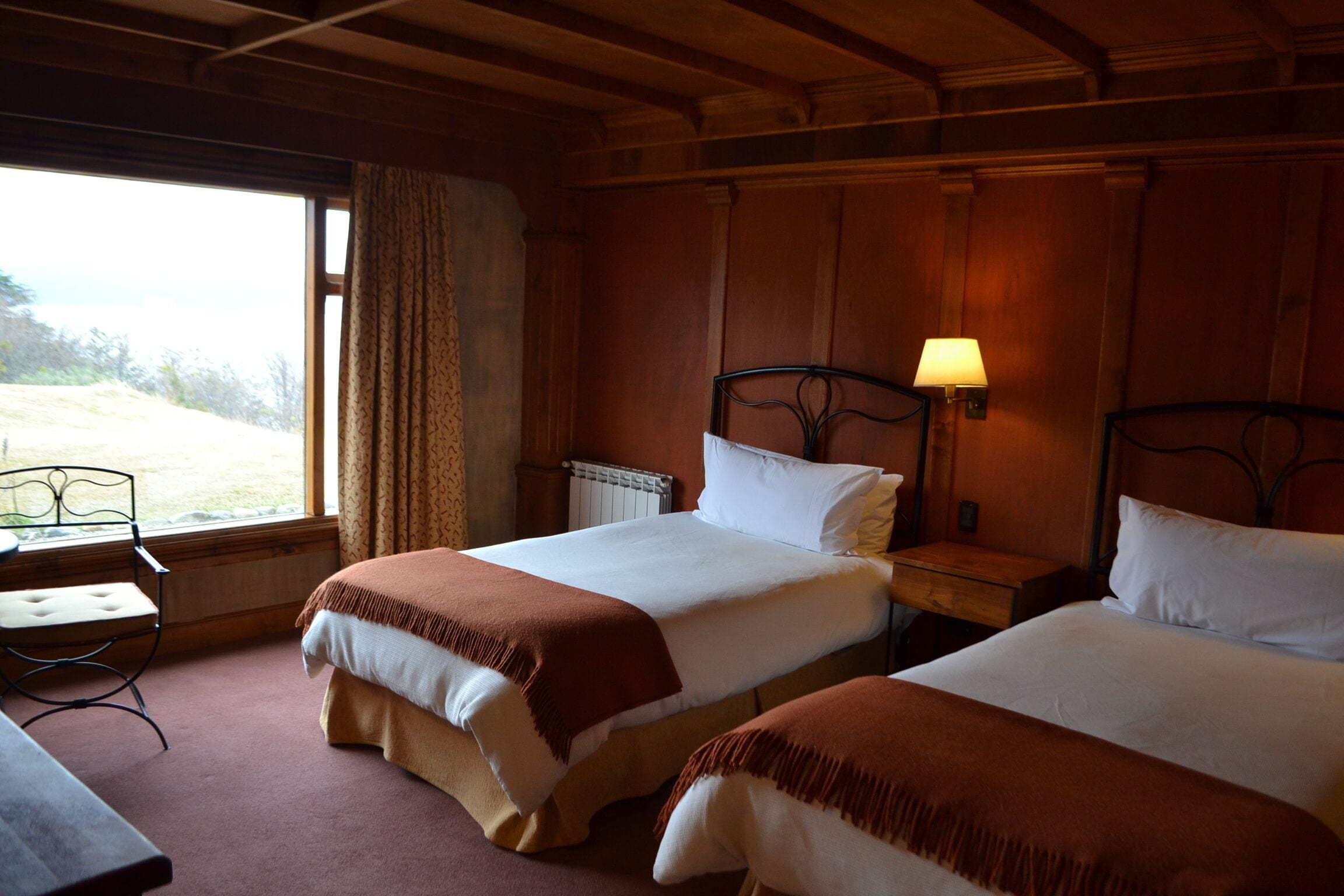 A hotel room with two beds, a window with curtains, a bedside table with a lamp, and a chair.