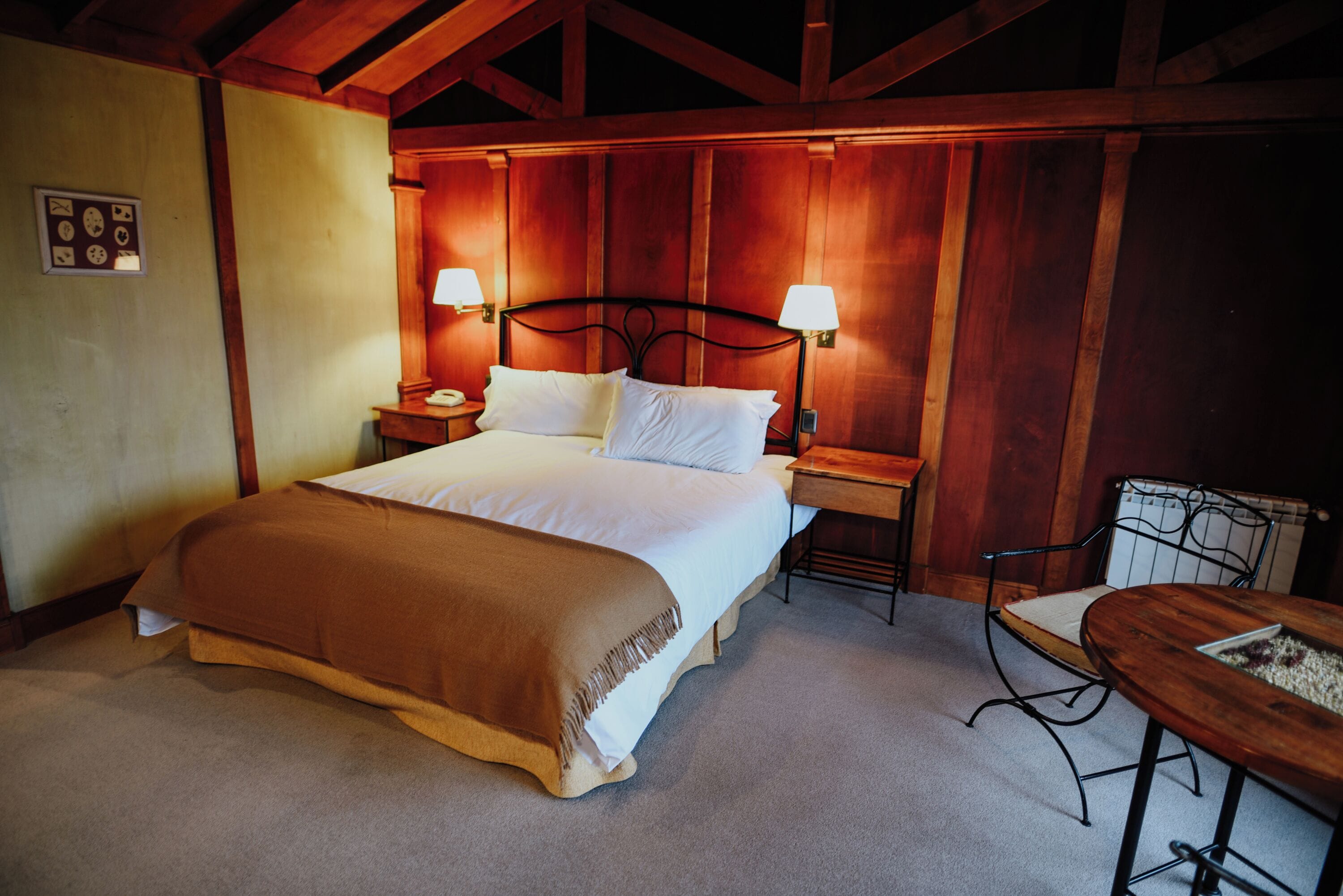 A neatly made bed with a brown blanket, two bedside lamps, a nightstand, and a small wooden table with a chair.