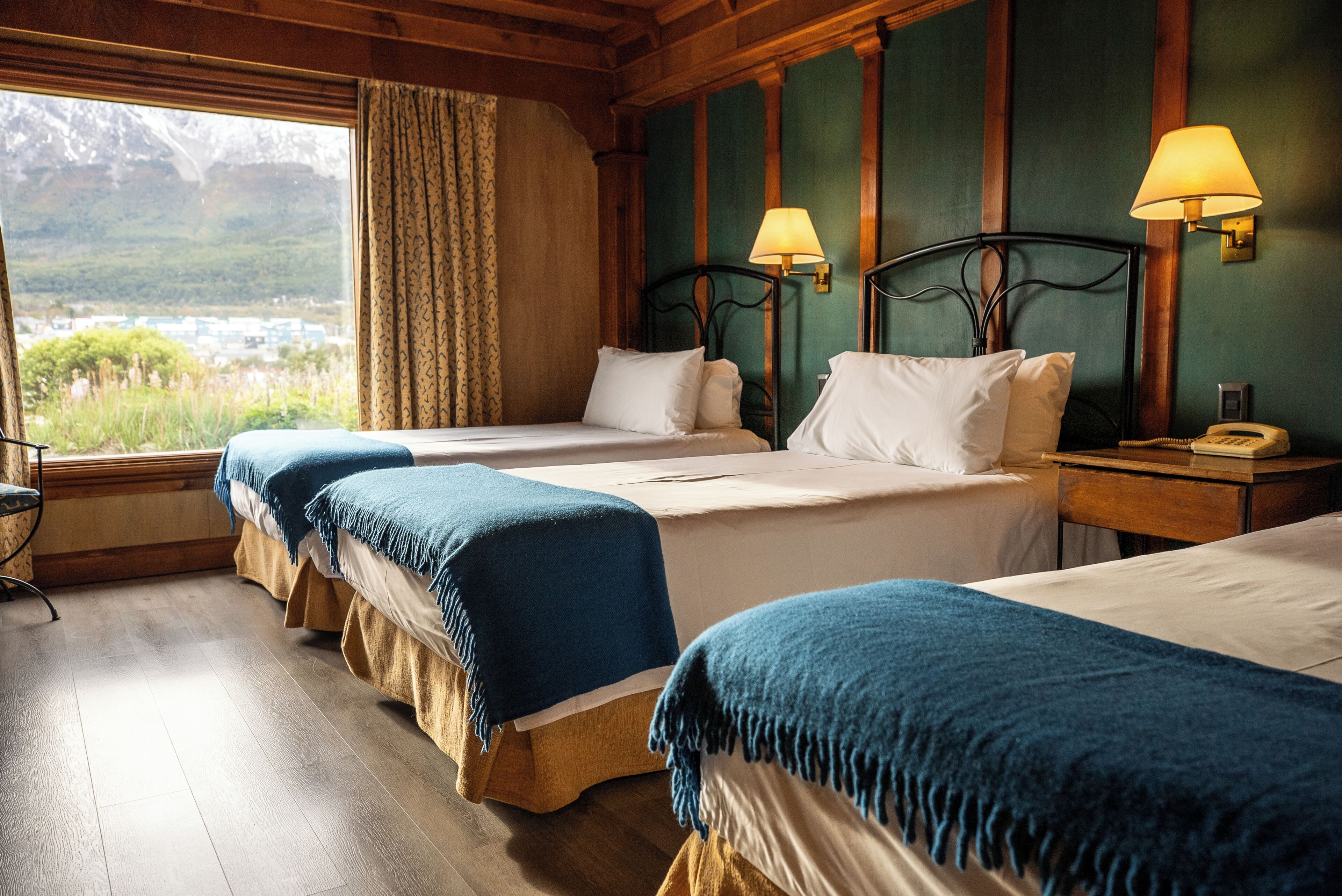 Habitación de hotel con tres camas, suelo de madera, vista al exterior y un teléfono en la mesita de noche.