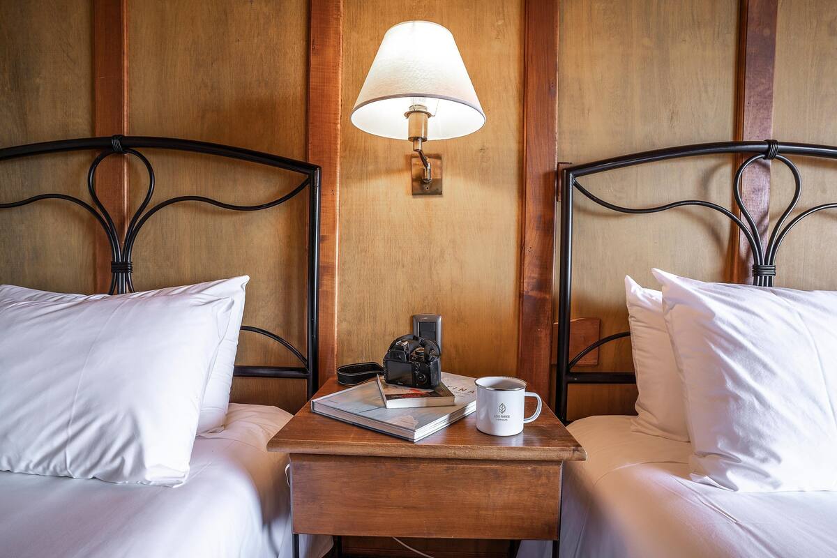 Et hotelværelse med to senge, et natbord med lampe, et kamera, en bog og en kaffekop.