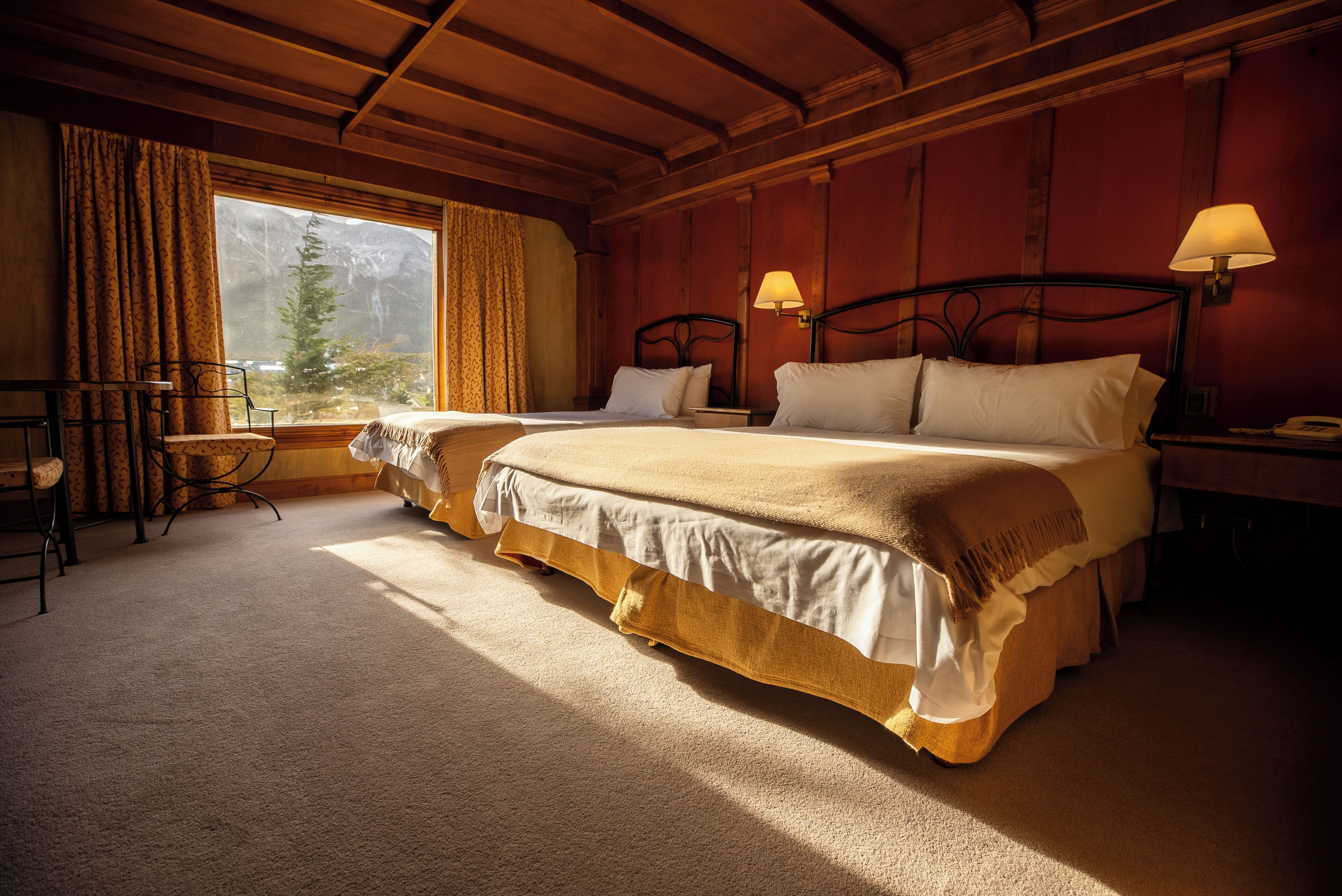 Una habitación de hotel con dos camas, techo de madera, ventana con cortinas, una mesita y una lámpara.