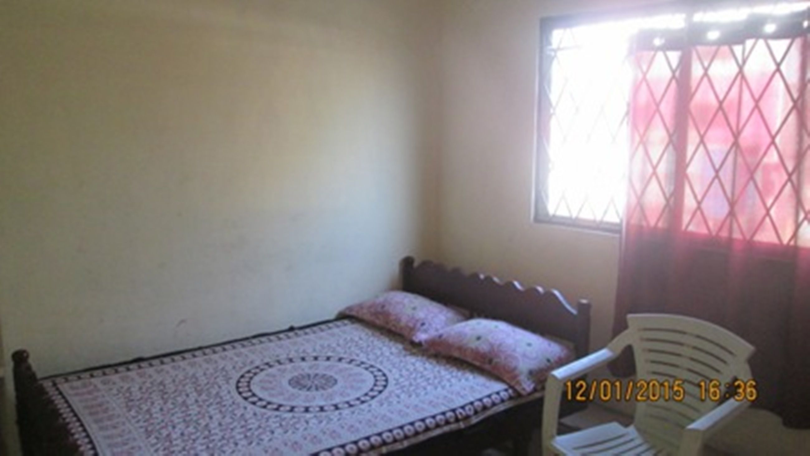 A small, single-bed room with a patterned bedspread, a wooden headboard, and a window with a metal grid.