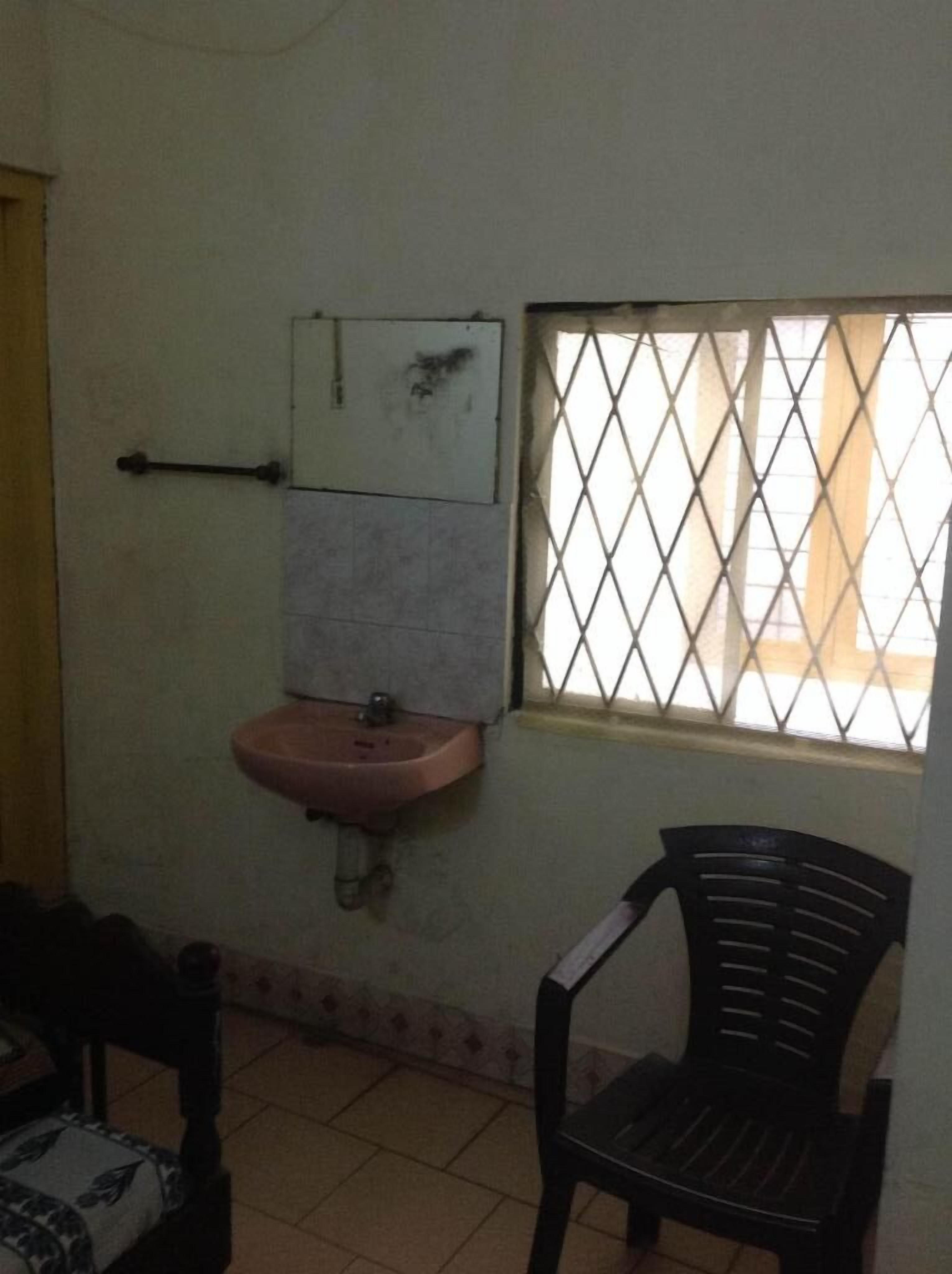 A small room with a pink sink, a window with a diamond-patterned blind, and a black plastic chair.