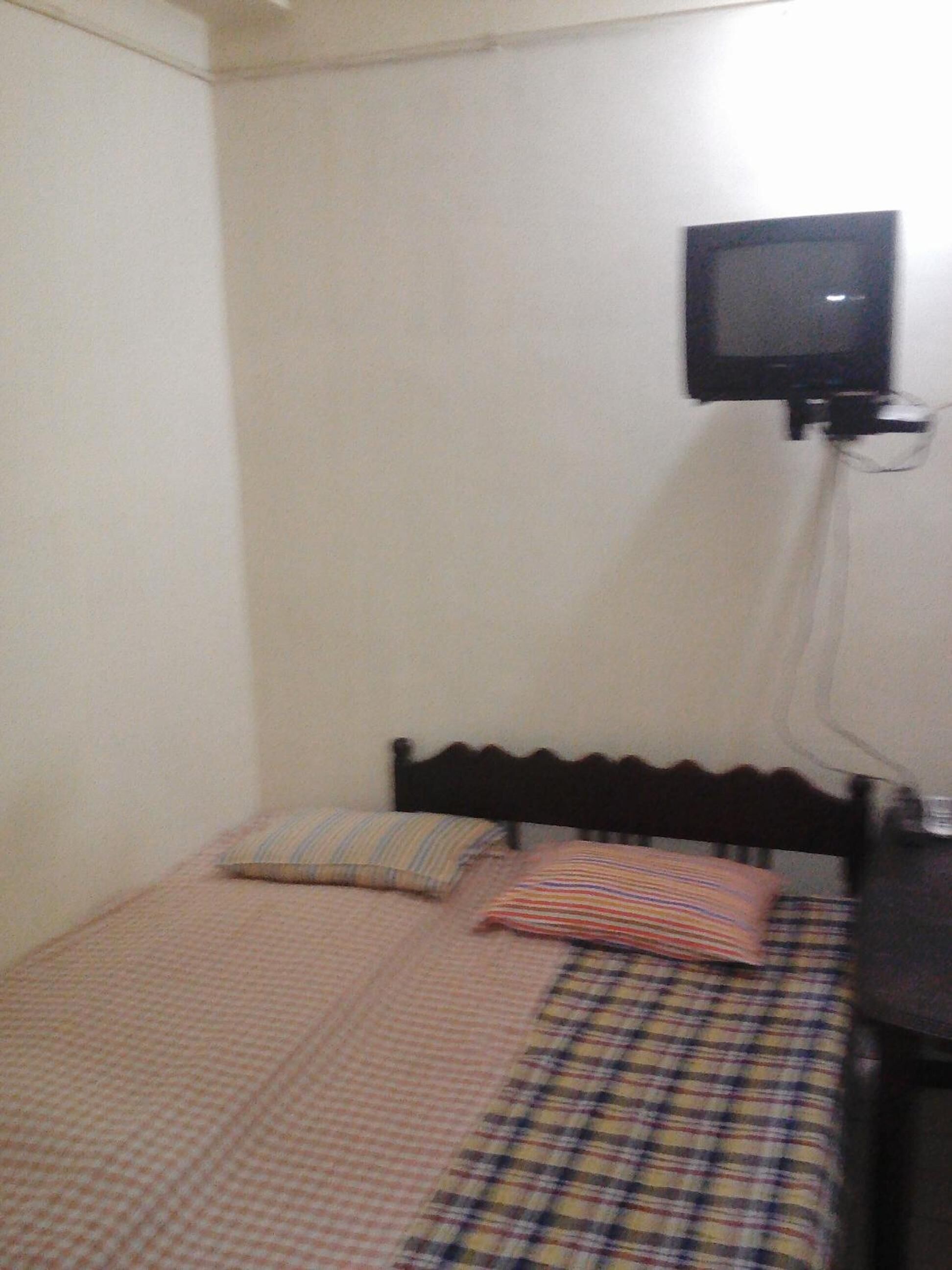 A small, simple room with a single bed, a television mounted on the wall, and a bedside table.