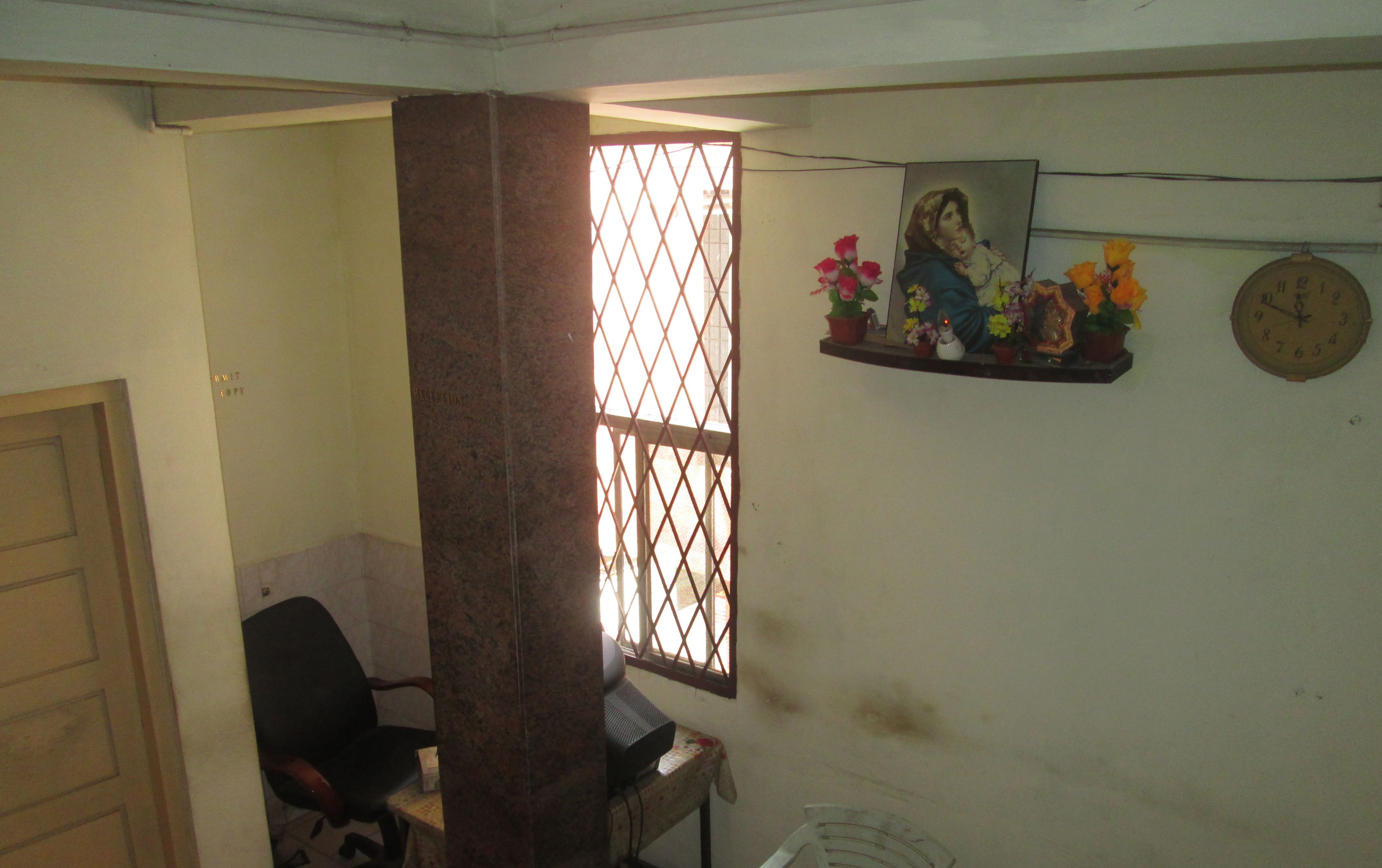 A room with a desk, chair, window with bars, a clock, and a shelf with flowers.