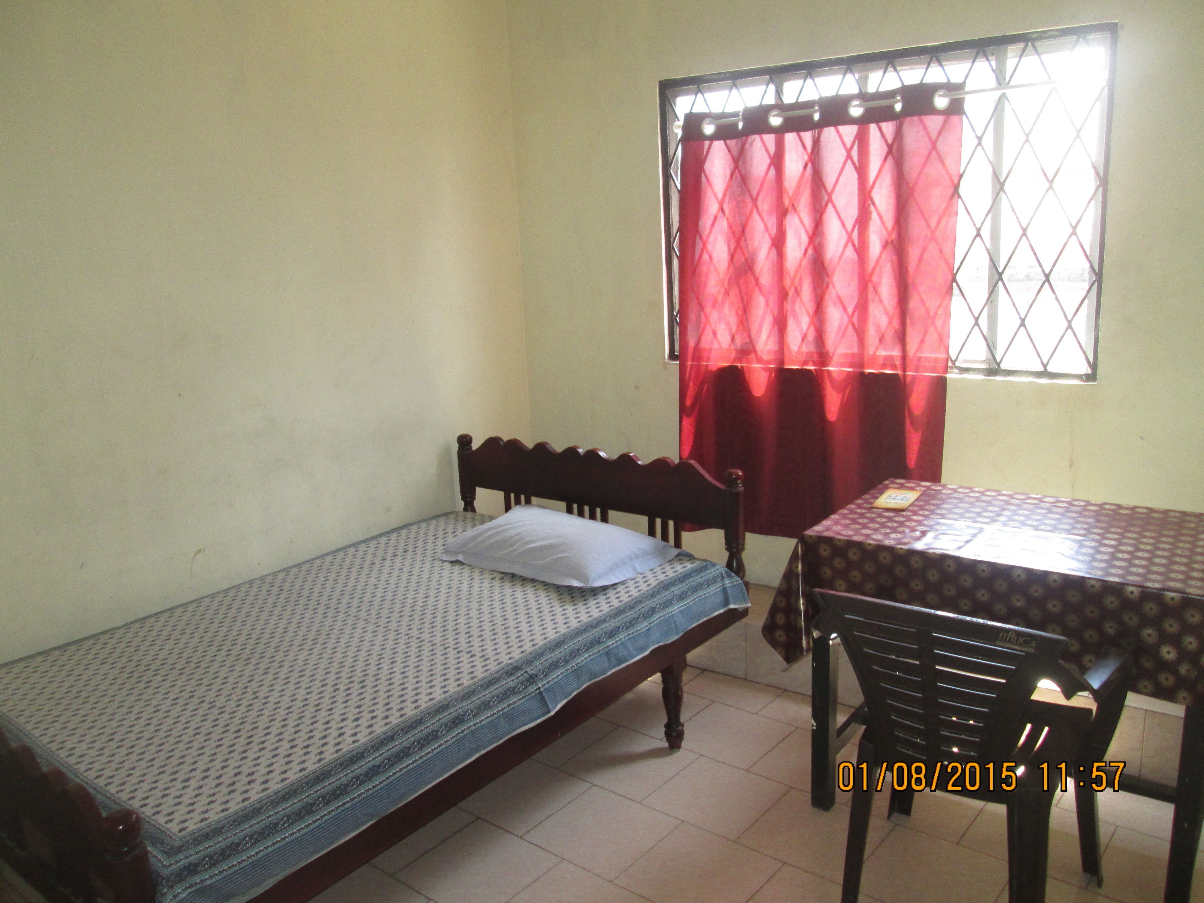 Une petite pièce avec un lit simple, une table et une fenêtre ornée d’un rideau rouge.