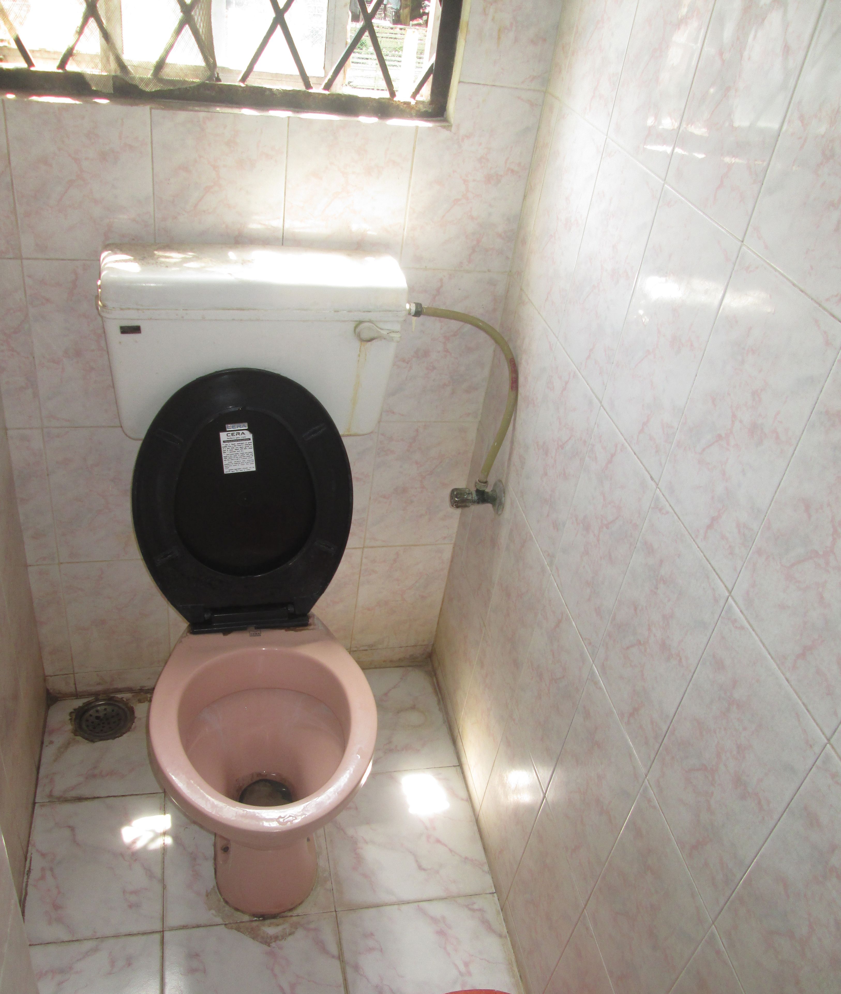 A small bathroom with a toilet, a visible water pipe, and a window with bars.