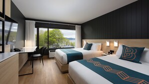 A modern hotel room with two beds, a desk, and a large window offering a view of trees and mountains. - Hôtel Baker (Gaspé)