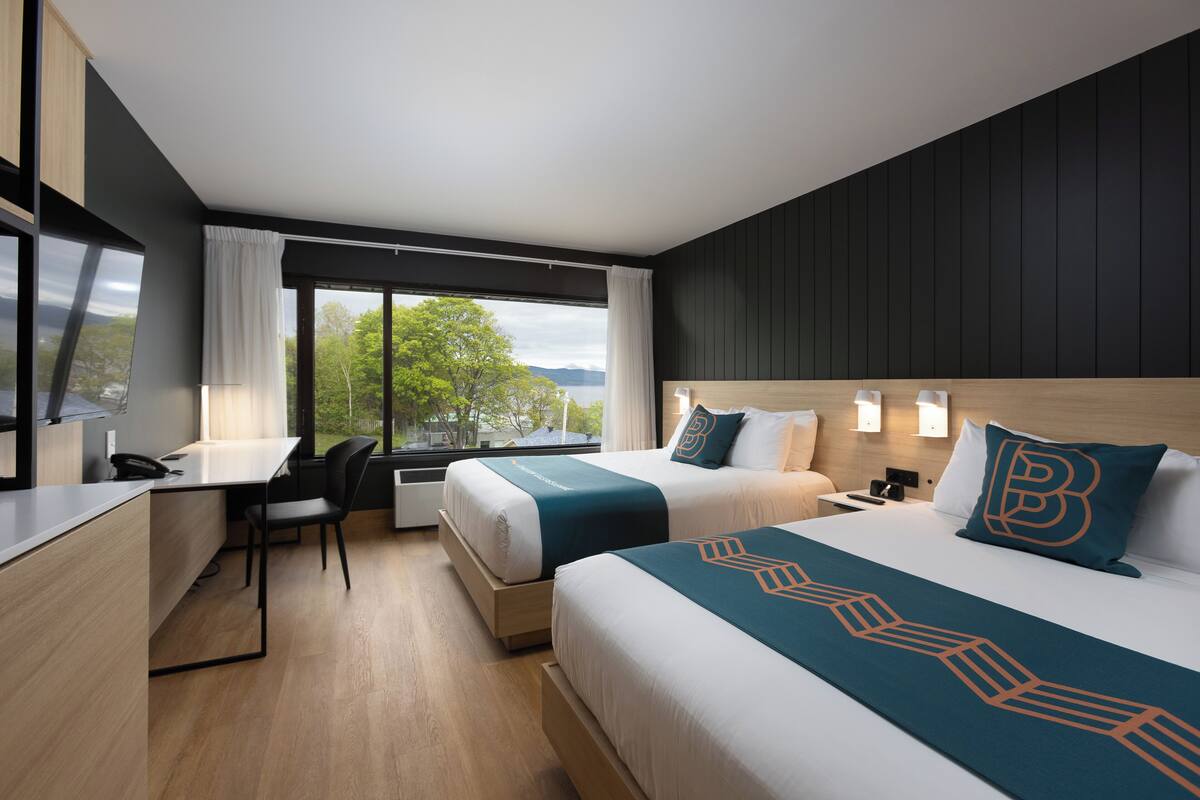 Ein modernes Hotelzimmer mit zwei Betten, einem Schreibtisch und einem großen Fenster mit Blick auf Bäume und Berge.