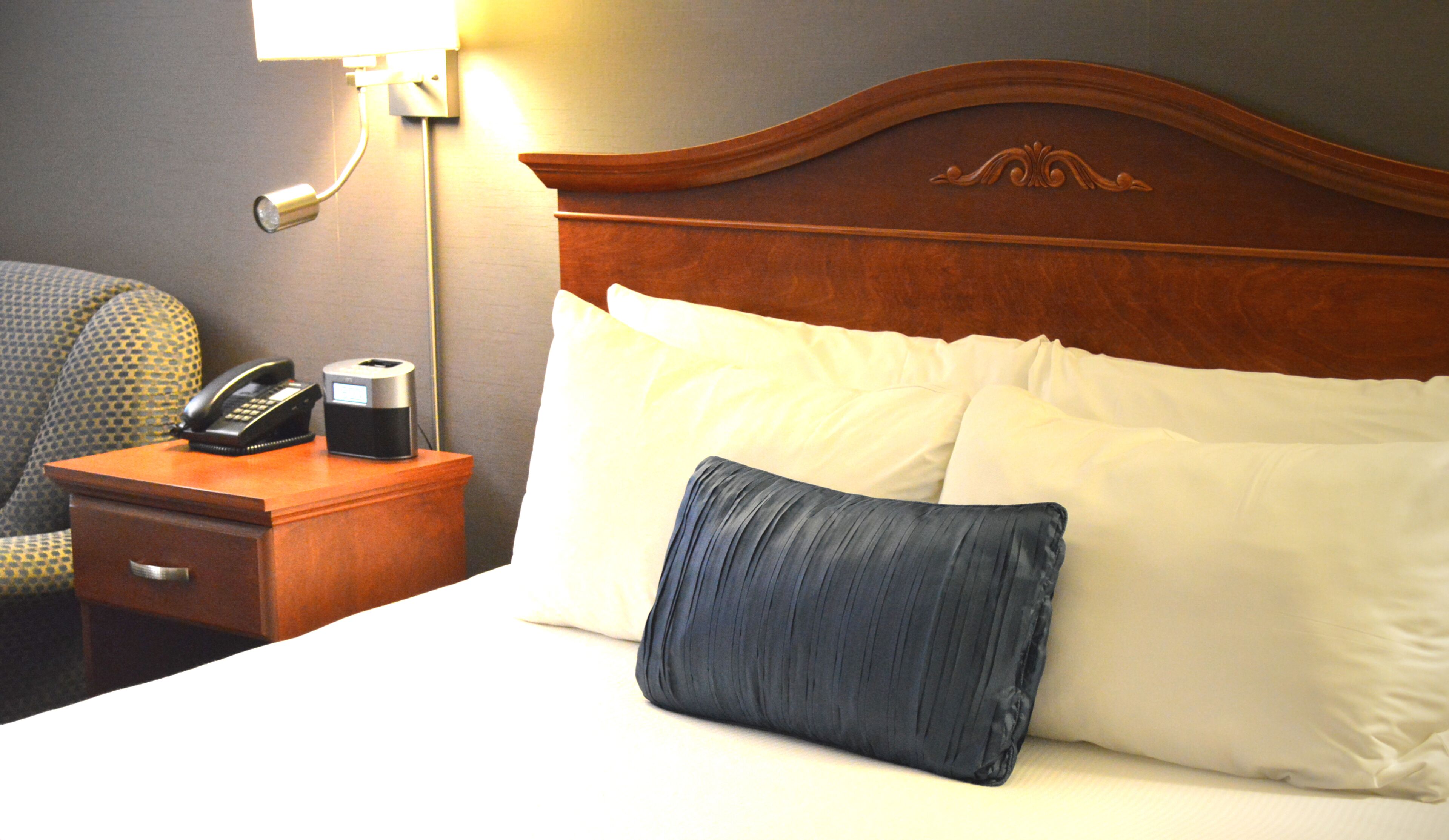 Ein Hotelzimmer mit einem Holzkopfteil, einem Nachttisch mit Telefon und Kaffeemaschine, einem Sessel und einem Bett mit weißen und blauen Kissen.