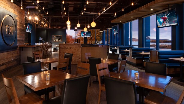 A restaurant with wooden tables and chairs, hanging pendant lights, and large windows with a view of the outdoors. - Hôtel Baker (Gaspé)