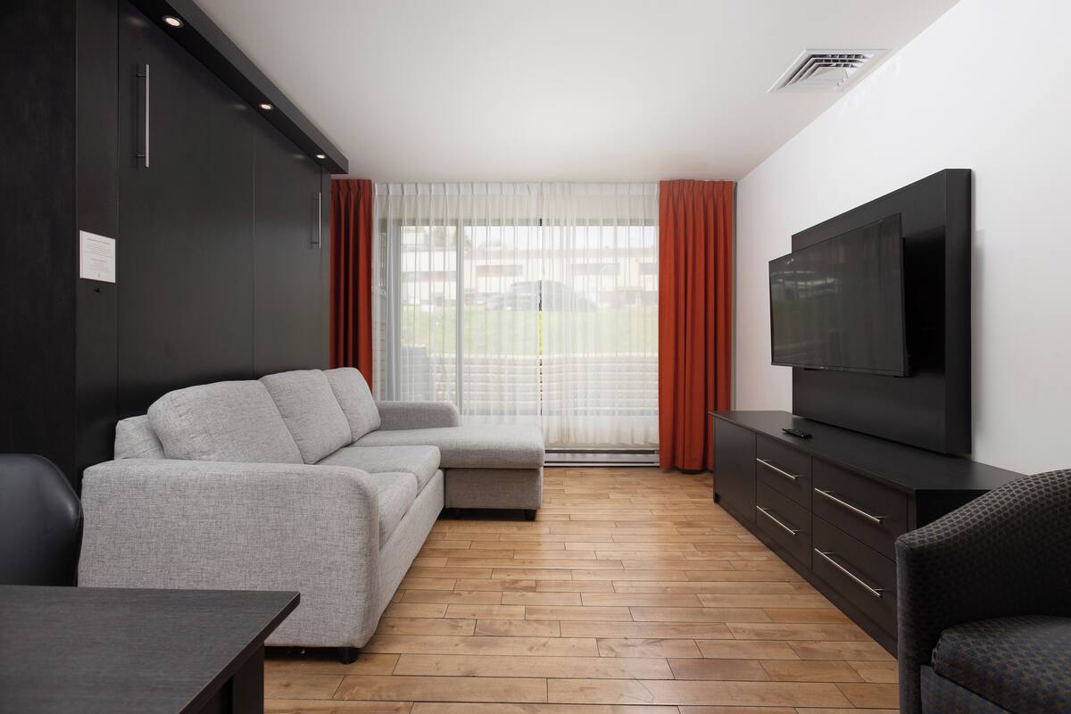 Ein modernes Wohnzimmer mit einem grauen Sofa, einem Fernseher auf einem schwarzen Schrank und einem Holzboden.