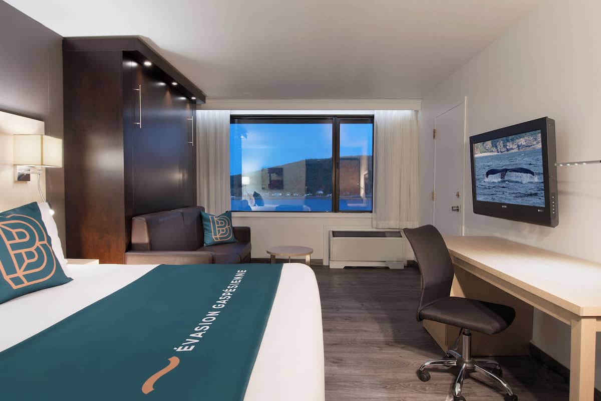 Ein Hotelzimmer mit Bett, Schreibtisch, Stuhl, Fernseher und Fenster mit Blick auf ein Gebäude und ein Gewässer.