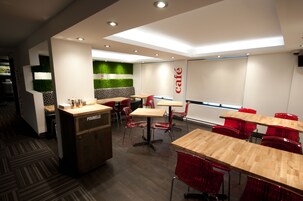 Interior de café moderno com mesas de madeira e cadeiras vermelhas, balcão com a etiqueta 'POUBELLE' e uma parede verde como detalhe decorativo.