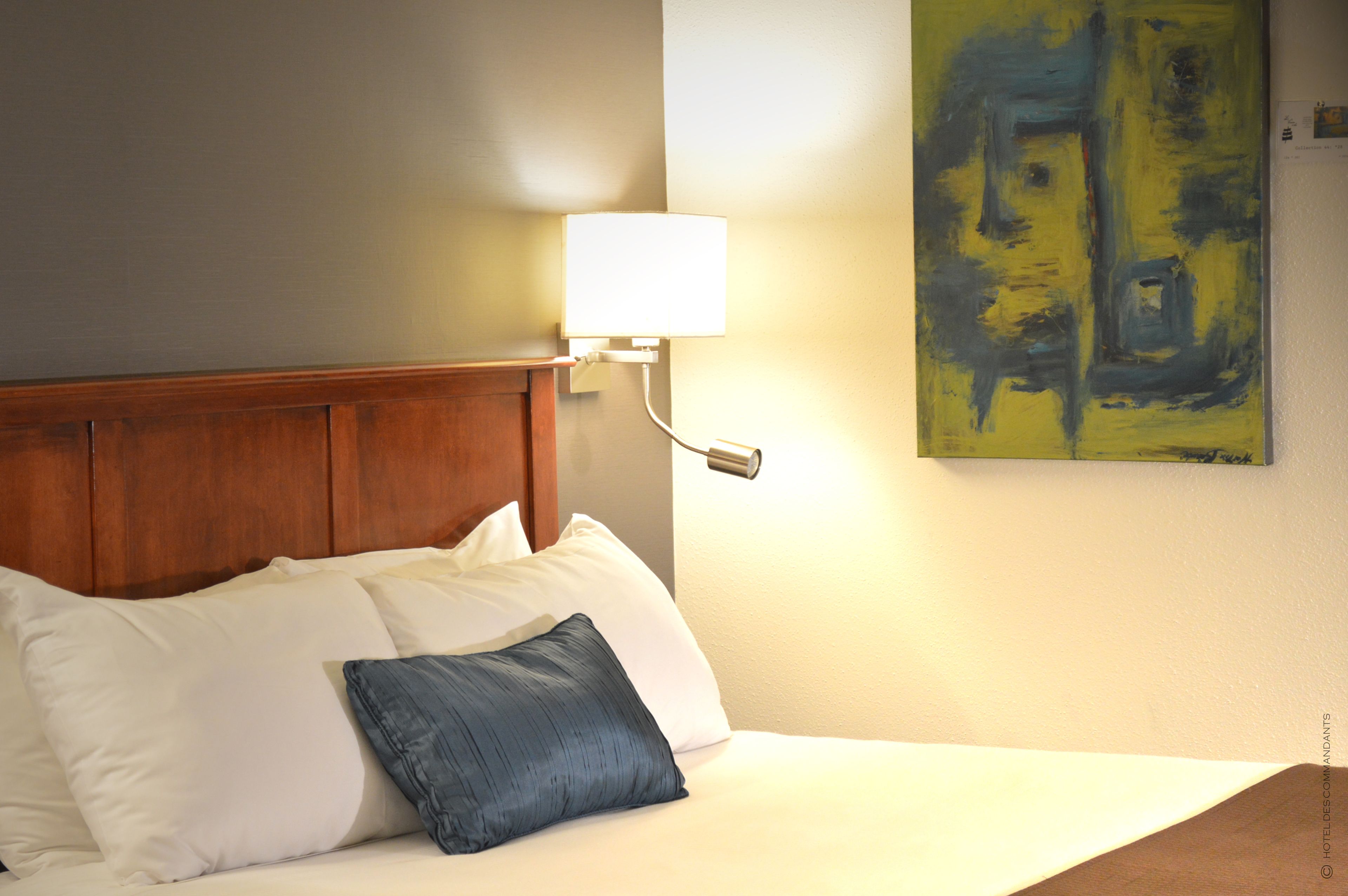 Habitación de hotel con cabecera de madera, cama con almohadas blancas y azules, lámpara de pared y una obra de arte abstracta.