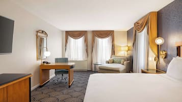 A hotel room with a large bed, a desk with a chair, a small table, a sofa, and a TV.