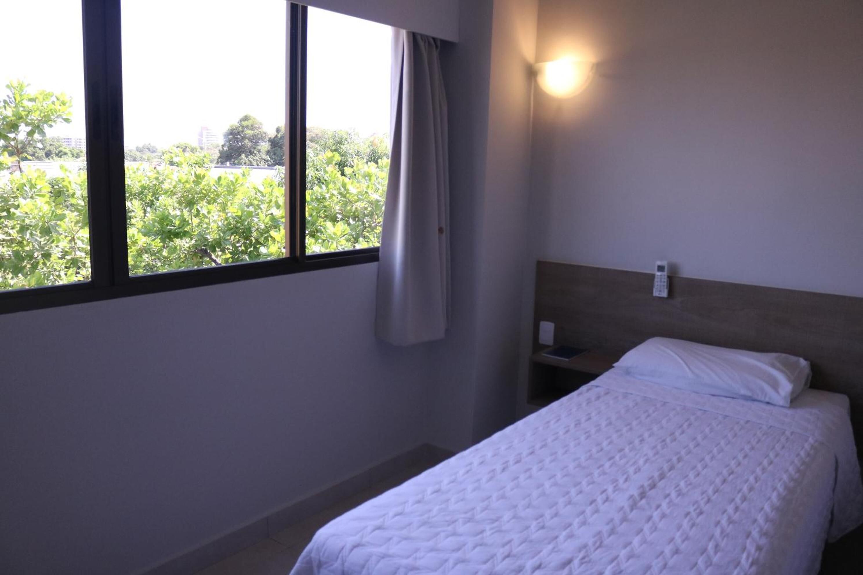 A hotel room with a bed, a nightstand, a wall-mounted phone, and a window with a view of greenery.