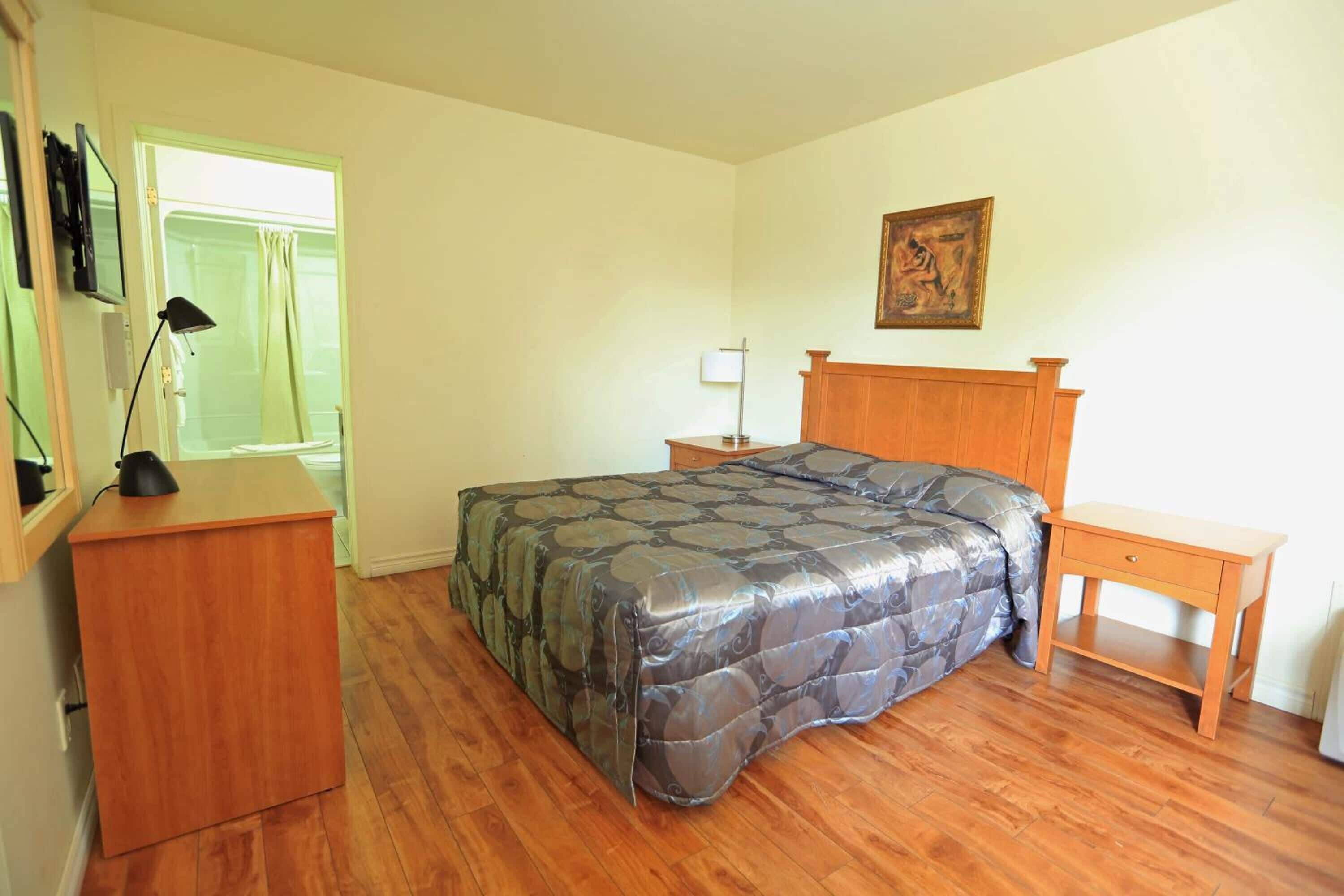 A bedroom with a bed, a wooden nightstand, a desk, a lamp, and a painting on the wall.