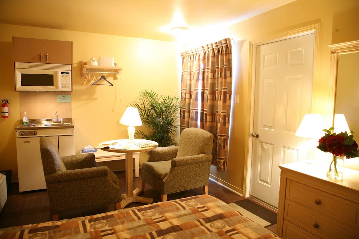 Habitación de hotel con cama, dos sillones, un escritorio pequeño con lámpara, microondas, lavamanos y puerta con espejo.
