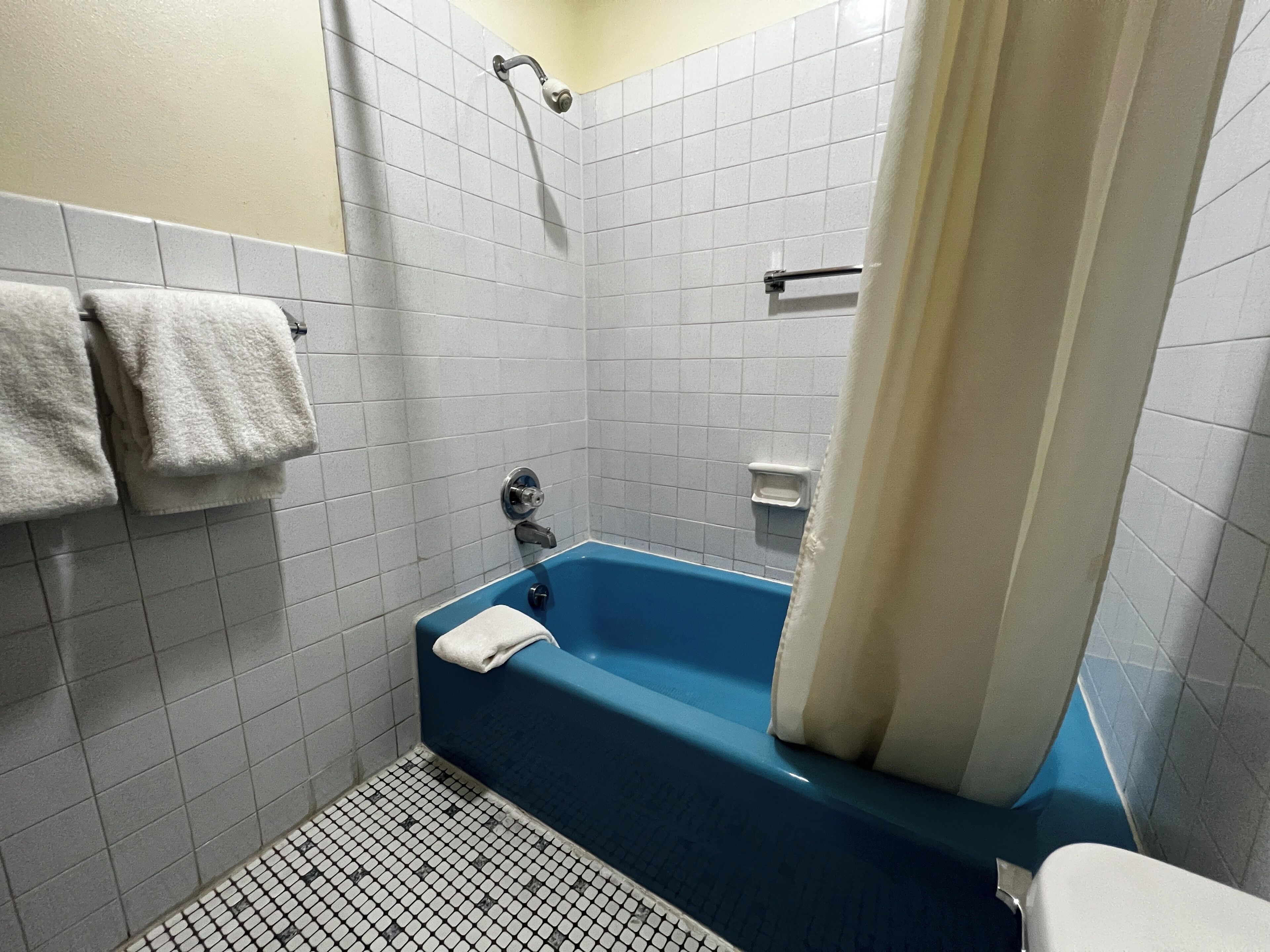 Un baño con una bañera azul, paredes cubiertas de azulejos blancos y un cabezal de ducha. Hay dos toallas colgadas en la pared.