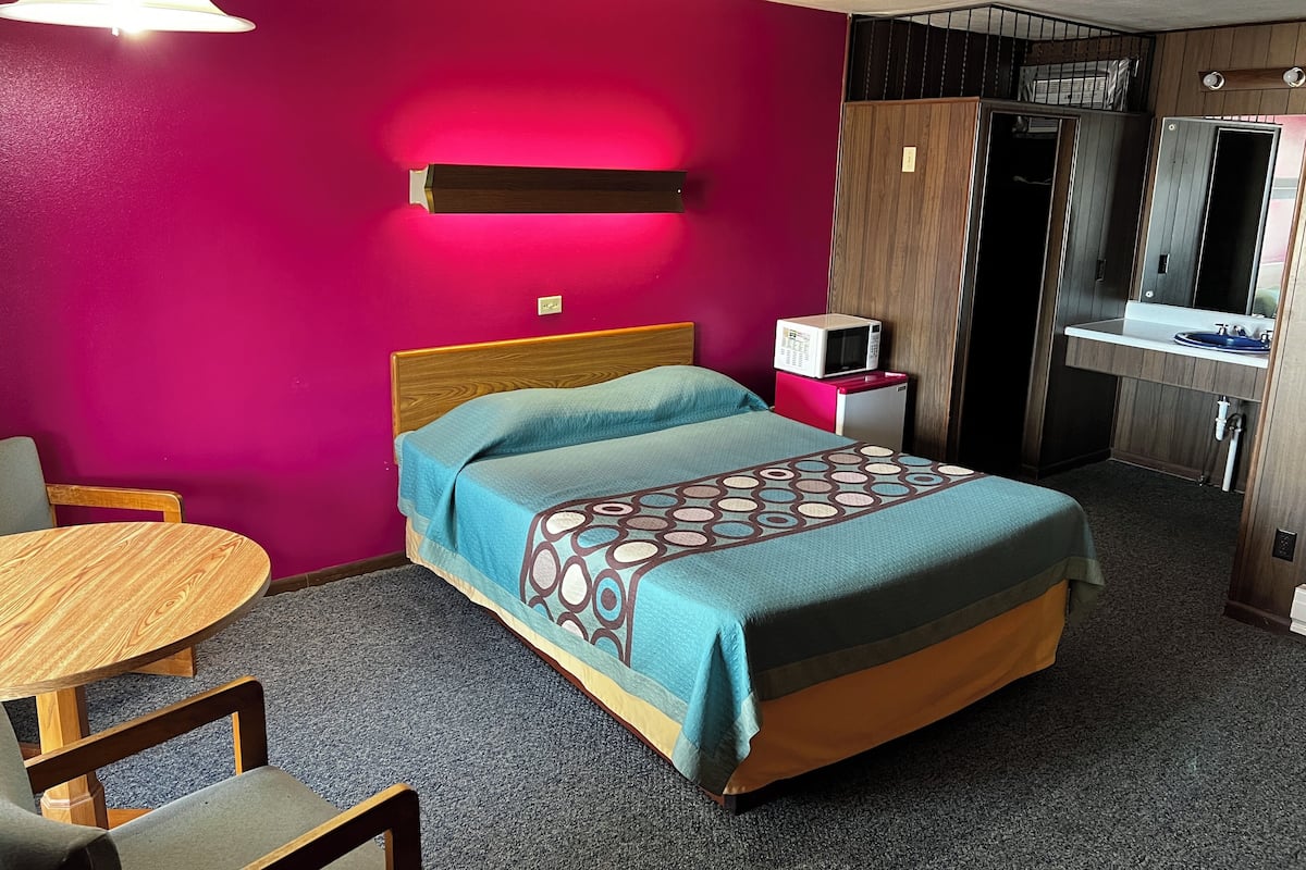 Quarto de hotel com cama, uma mesinha, uma cadeira, micro-ondas e pia.