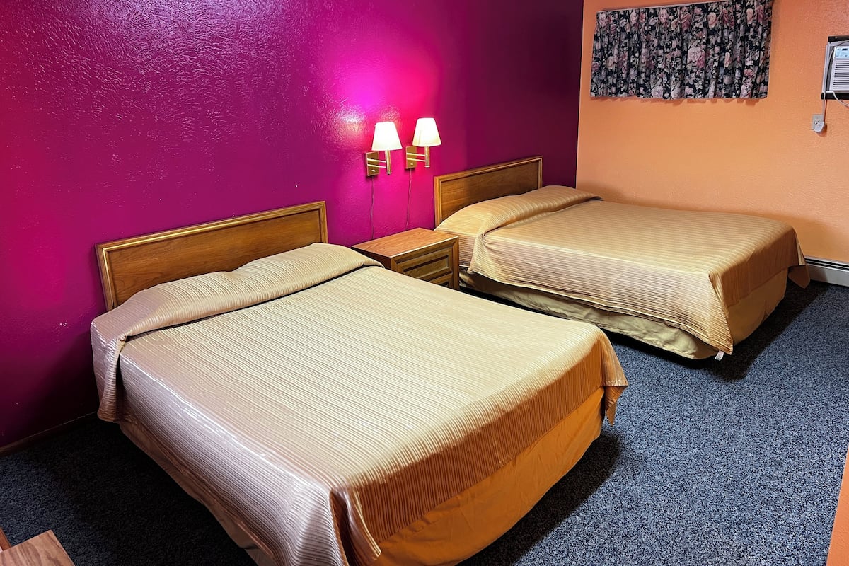 Quarto de hotel com duas camas, uma mesa de cabeceira, uma luminária na parede e cortina florida.
