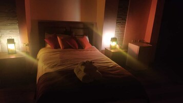 Un dormitorio con poca luz, una cama, almohadas y una mesita de noche con una lámpara.