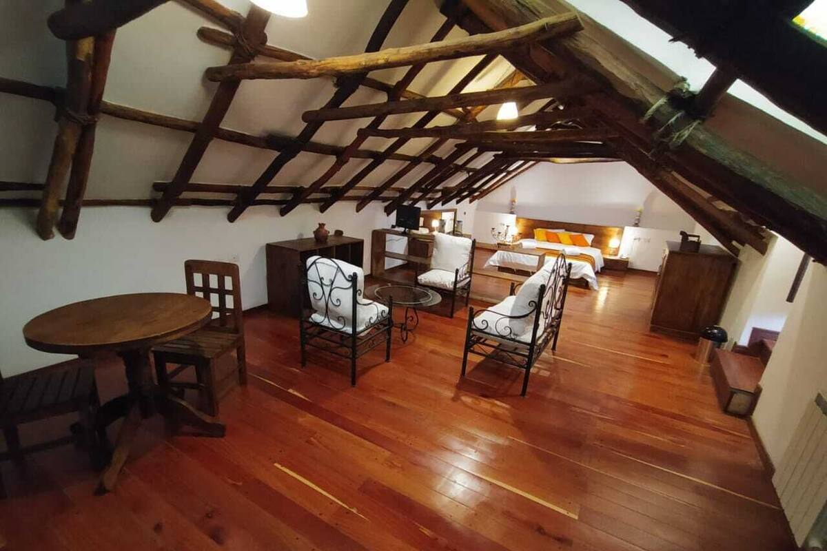 A rustic attic room with wooden beams, a dining table, four chairs, and a kitchen area in the background.