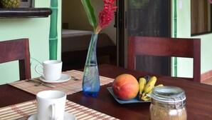 Una mesa de comedor con una taza blanca, un jarrón azul con una flor roja, un plato con mango y plátanos, y un tarro de azúcar.