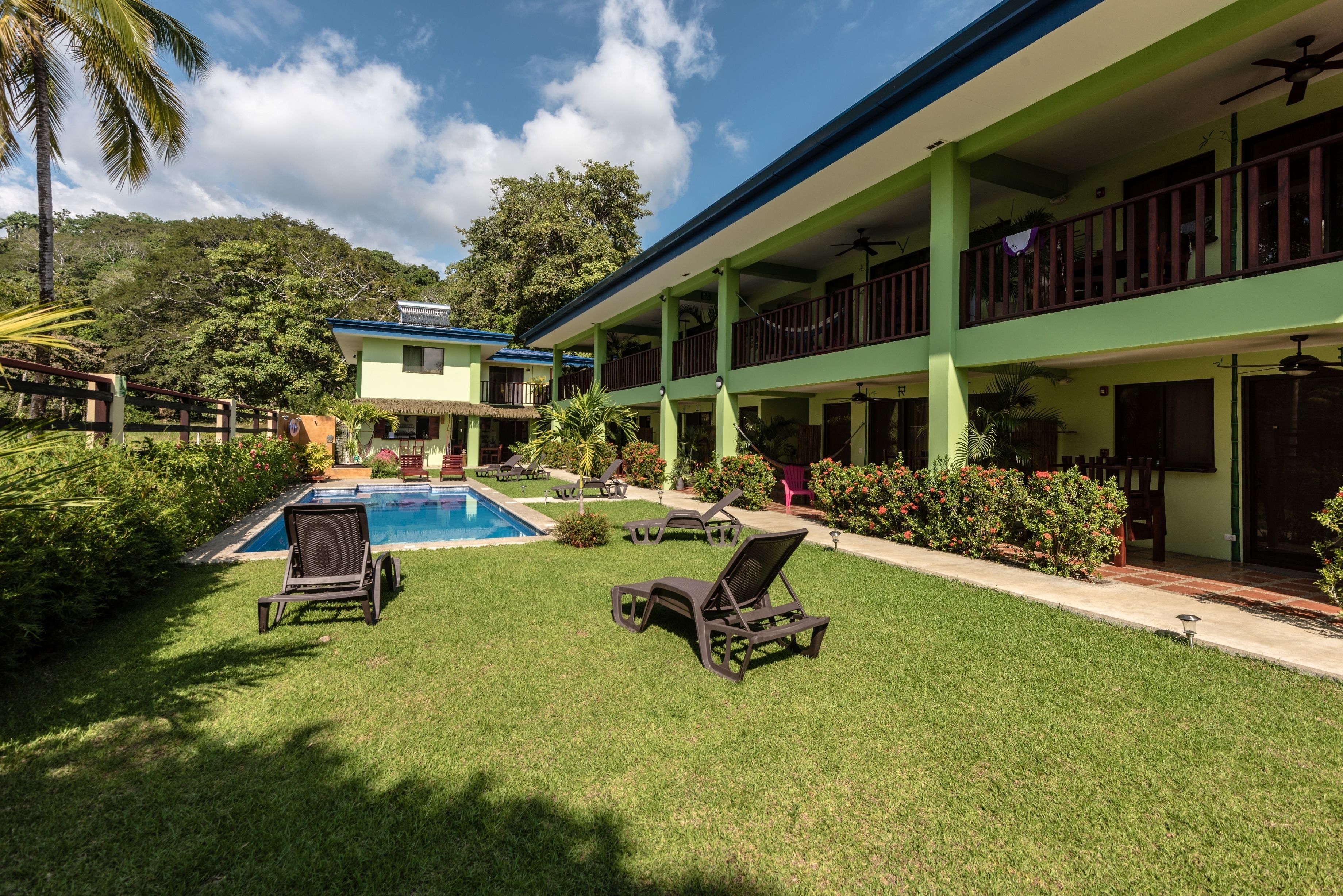 A hotel with a pool, two lounge chairs, and a green building with balconies.