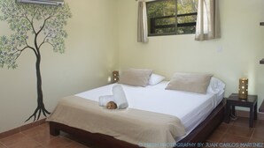 Un dormitorio con una cama, un ventilador de techo, un equipo de aire acondicionado, una ventana con cortinas y un mural en la pared de un árbol.