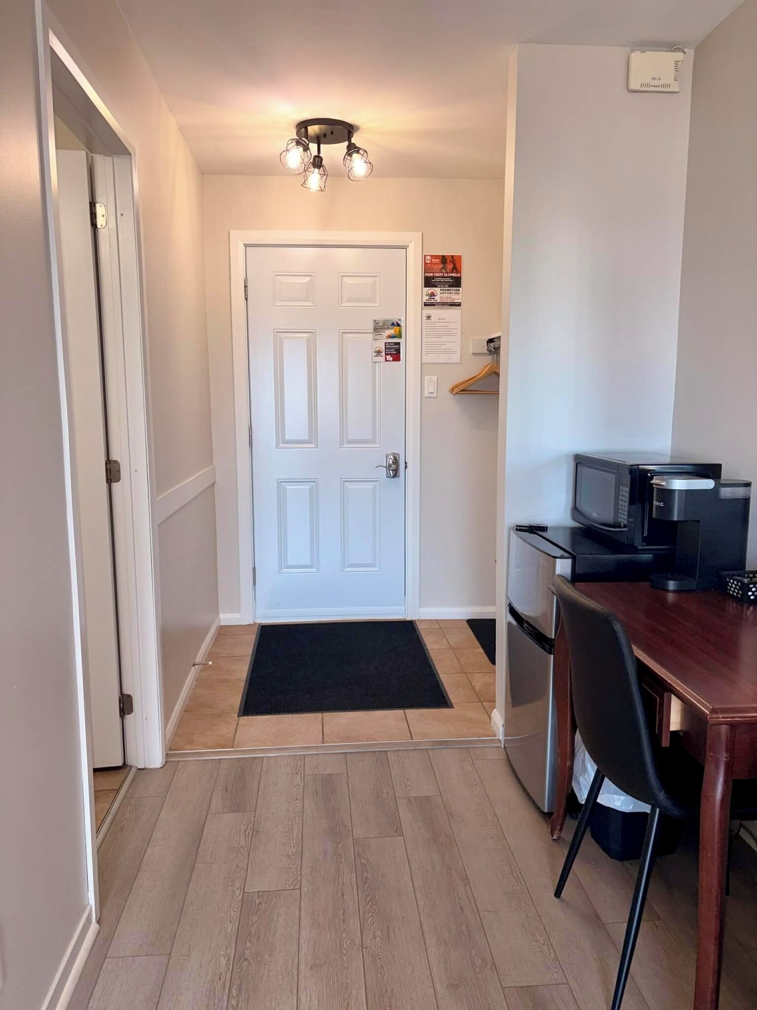 A narrow hallway with a white door, a small kitchenette with a microwave, and a wooden dining table with a chair.