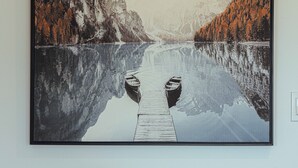 Een lijst met een landschap waarin een meer, boten, een steiger en bergen zijn afgebeeld. Daaronder staat een ronde tafel met een bloempot.