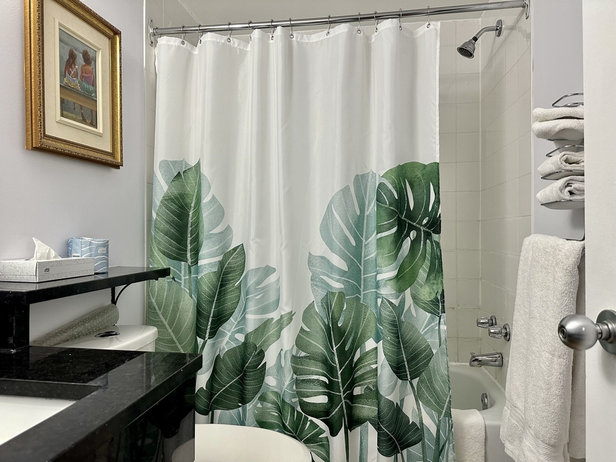 A bathroom with a shower curtain featuring a tropical leaf pattern, a framed picture on the wall, and a towel rack with folded towels.