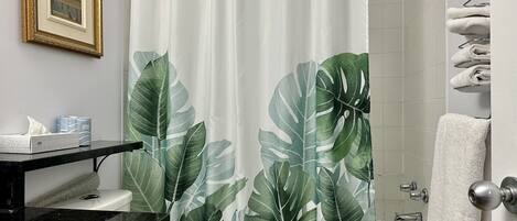 A bathroom with a shower curtain featuring a tropical leaf pattern, a framed picture on the wall, and a towel rack with folded towels.