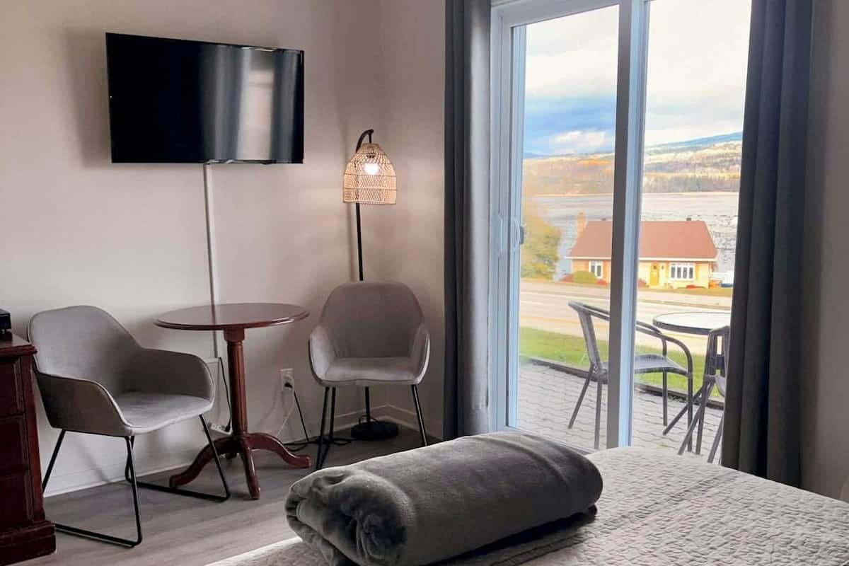 Habitación de hotel con una cama grande, dos sillones, una mesita, un televisor y vista a una casa por la ventana.