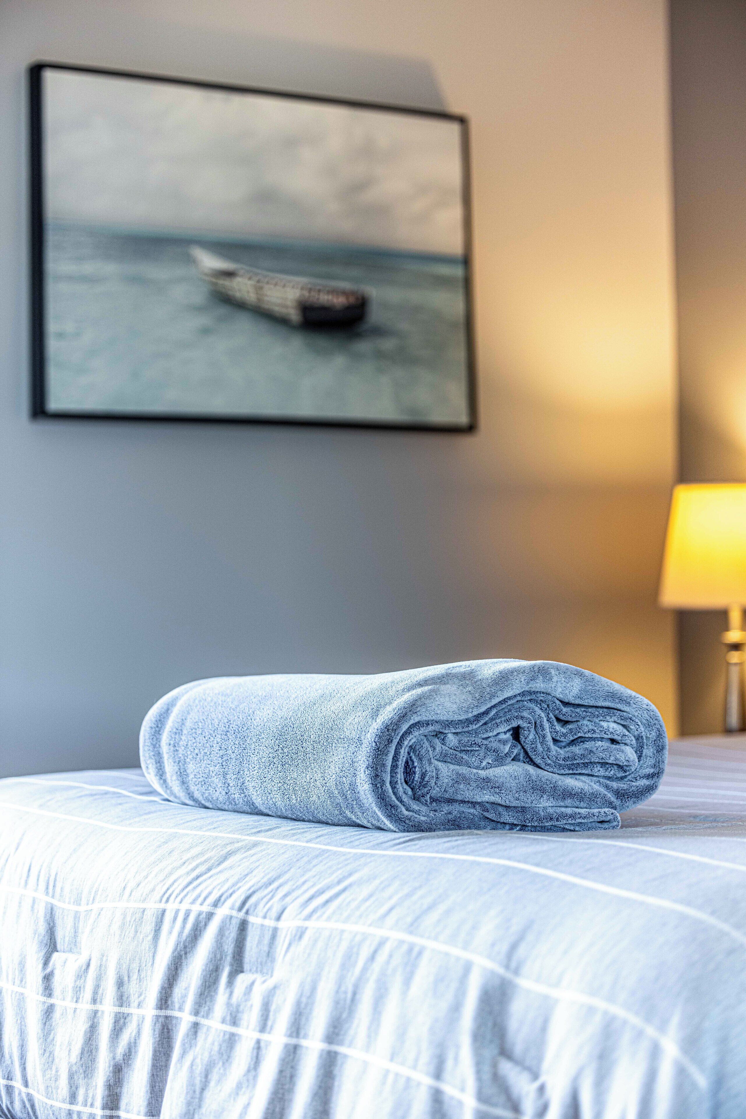 Een netjes gevouwen handdoek op een bed met een lichtblauwe en witte strepen dekbedovertrek, een lijst met een foto van een boot op het water aan de muur, en een lamp met een gele lampenkap aan de rechterkant.