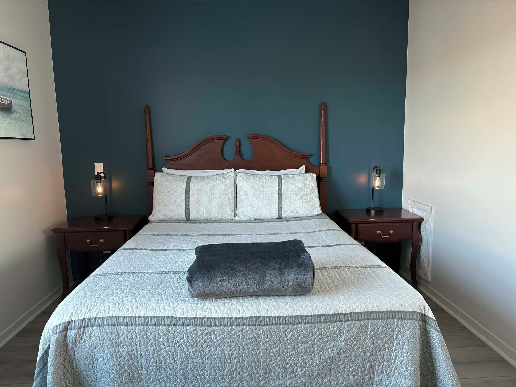 A neatly made bed with a grey blanket and pillows, two bedside tables with lamps, and a dark blue wall.