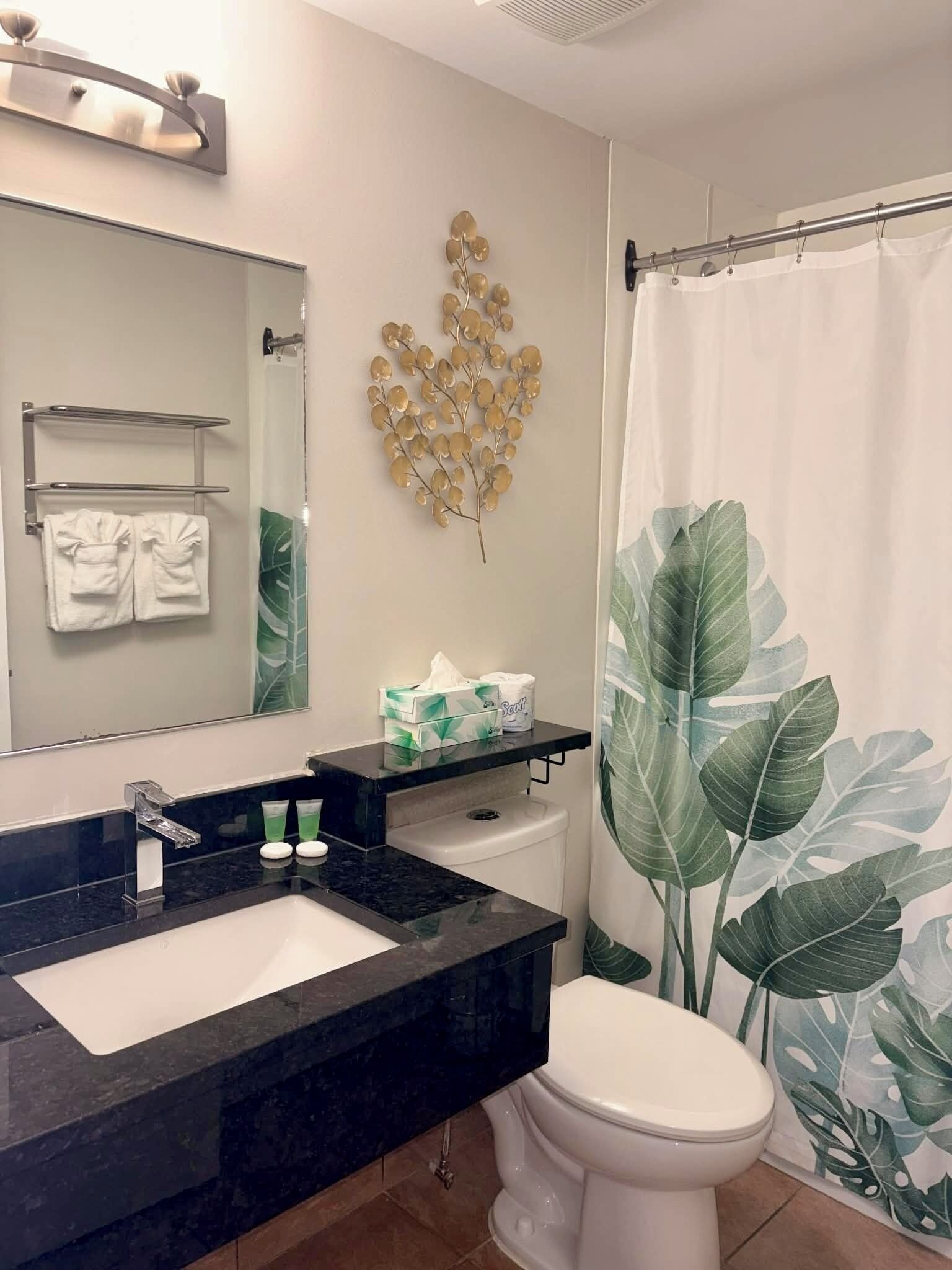 Une salle de bain avec un lavabo noir, des toilettes blanches et un rideau de douche orné d’un motif de feuilles.