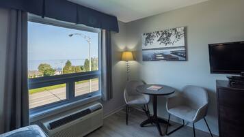 Habitación de hotel con un ventanal grande, una mesa con dos sillas, una lámpara, un televisor y un cuadro en la pared.